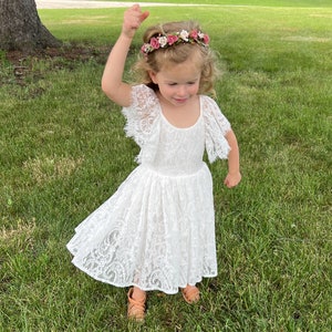 May include: A young girl wearing a white lace dress with a floral crown on her head. She is standing in a grassy area and smiling.