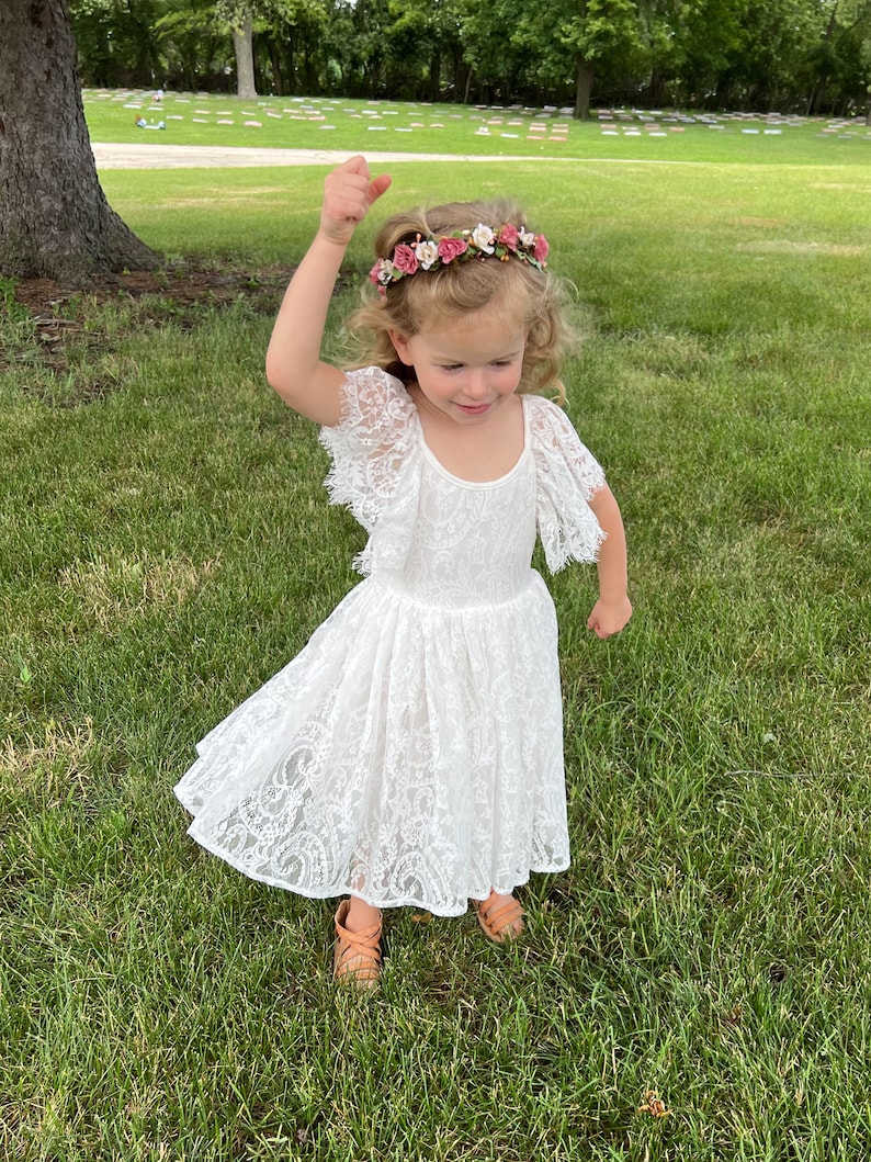 May include: A young girl wearing a white lace dress with a floral crown on her head. She is standing in a grassy area and smiling.