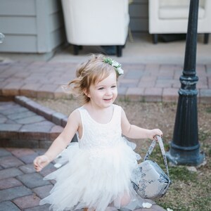 Op de afbeelding: Een jong meisje in een witte tule jurk en een bloemenkrans loopt over een geplaveid pad en draagt een mand met bloemenpatroon.