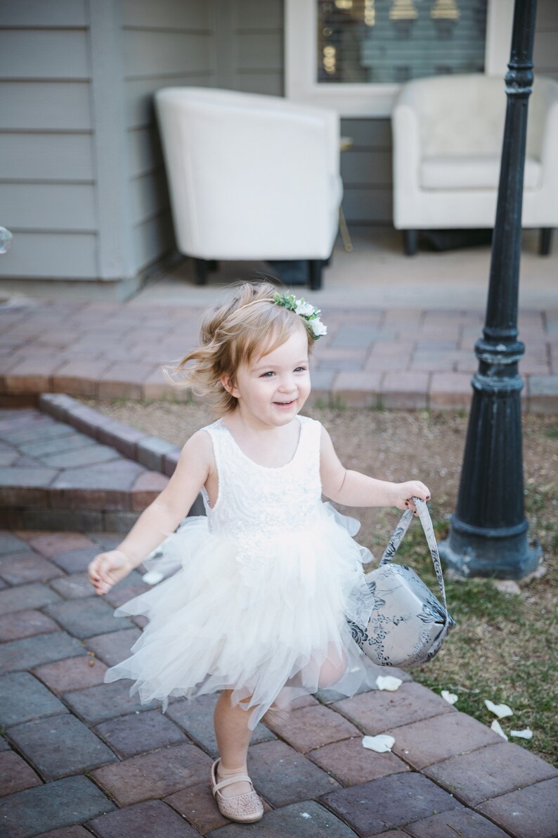 Op de afbeelding: Een jong meisje in een witte tule jurk en een bloemenkrans loopt over een geplaveid pad en draagt een mand met bloemenpatroon.