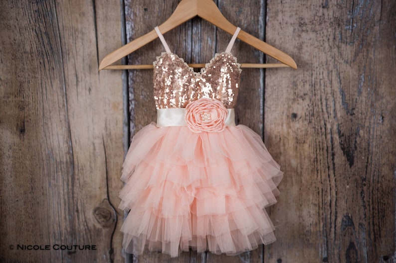 May include: A peach tulle dress with a rose gold sequin bodice and a satin sash with a large flower accent. The dress is hanging on a wooden hanger.