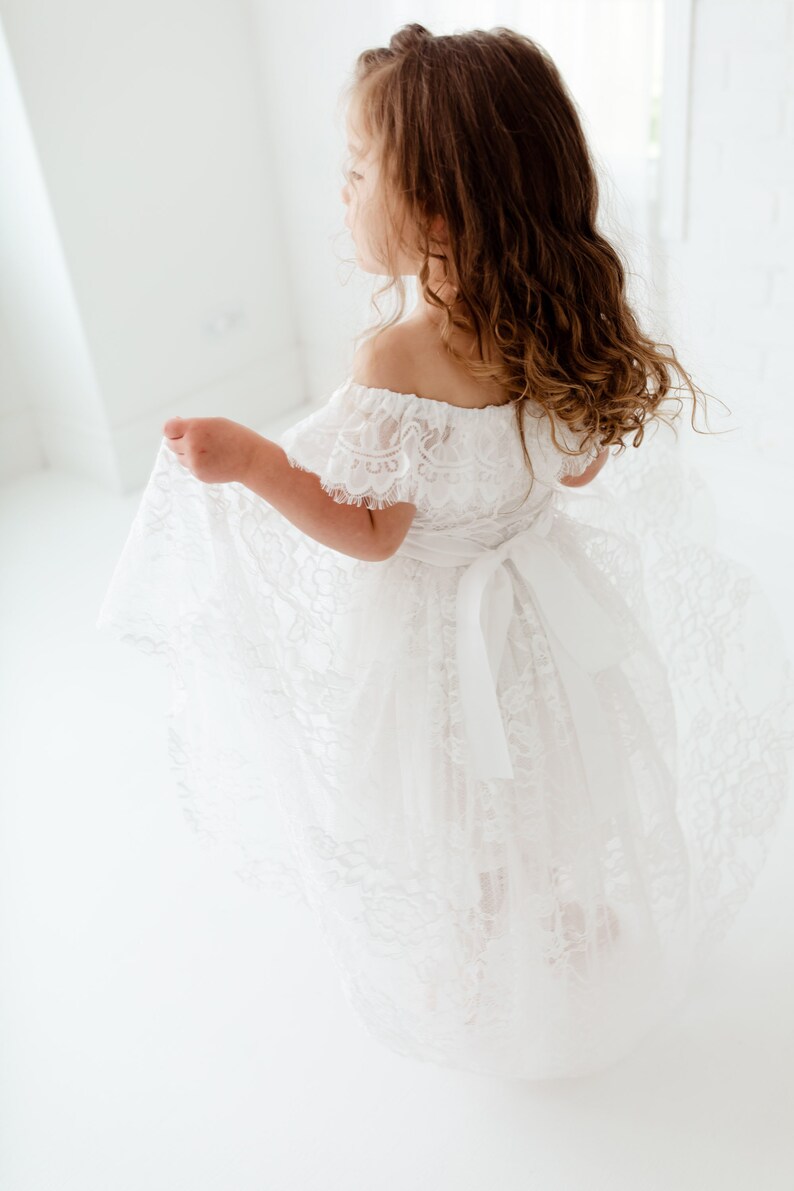 May include: A young girl wearing a white off-the-shoulder lace dress with a satin ribbon tied at the waist. The dress has a flowing skirt and is perfect for a special occasion.