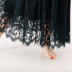 May include: A dark navy blue lace skirt featuring an intricate floral pattern throughout the fabric. The hemline is finished with a scalloped edge that displays delicate, fine fringe detailing along the bottom of the garment.