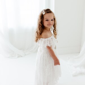 May include: A young girl with curly blonde hair wears a white off-the-shoulder lace dress with a pearl hair clip. She is standing on a white background.