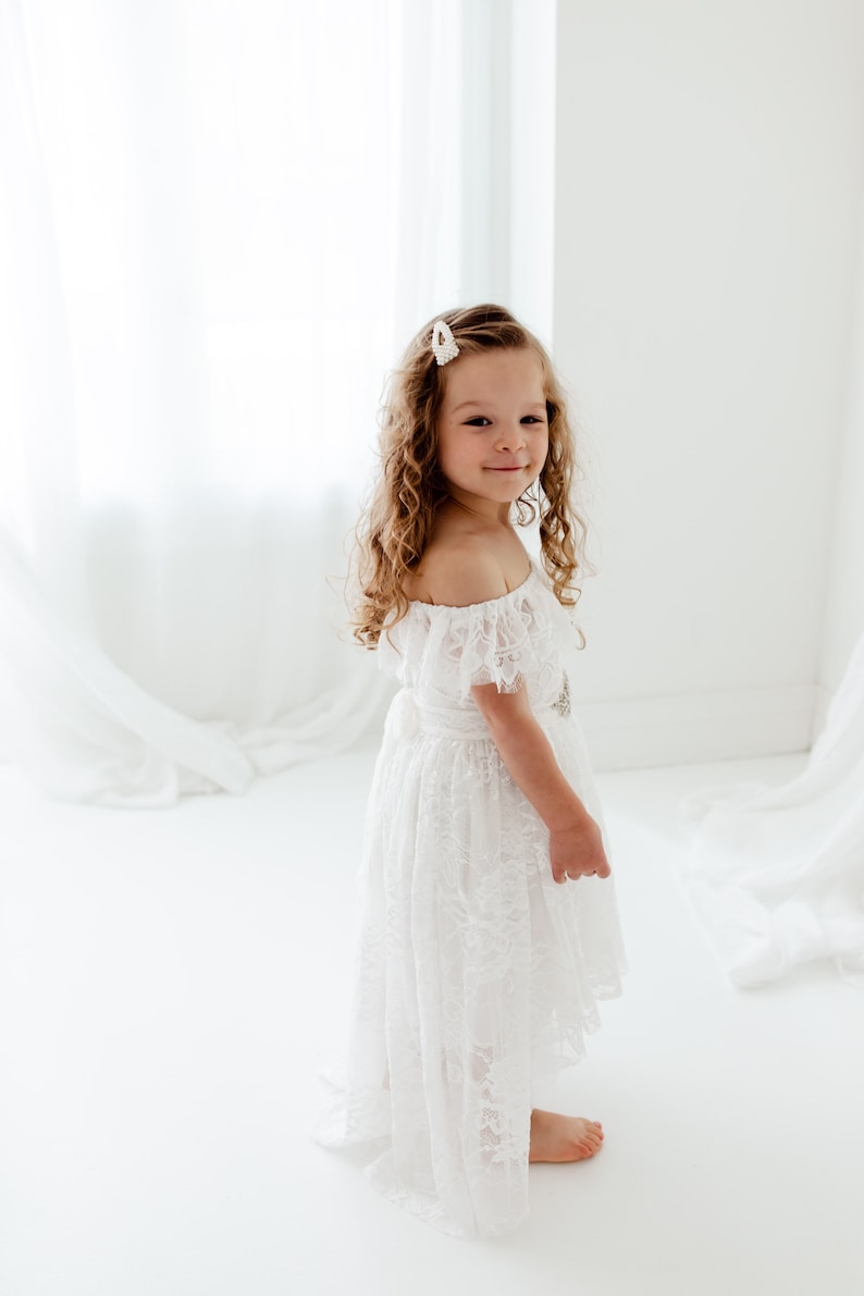 May include: A young girl with curly blonde hair wears a white off-the-shoulder lace dress with a pearl hair clip. She is standing on a white background.