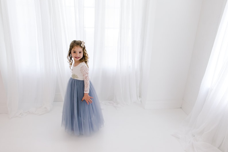 May include: A young girl wearing a white and blue tulle dress with lace sleeves. She is standing in front of a white background with white curtains.