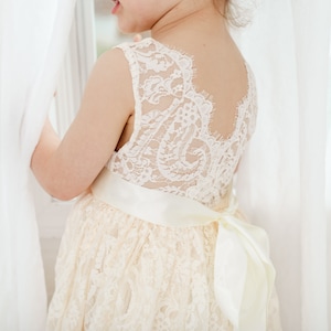 May include: A young girl wearing a white lace dress with a satin sash and bow. The dress has a scalloped neckline and a lace-up back.