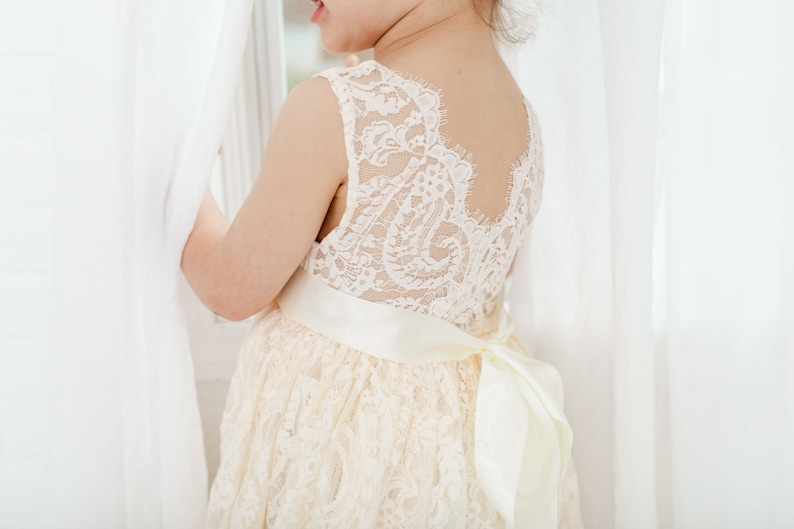 May include: A young girl wearing a white lace dress with a satin sash and bow. The dress has a scalloped neckline and a lace-up back.