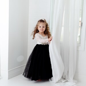 May include: A floor-length dress featuring a white long-sleeved lace bodice and a voluminous black tulle skirt. The waist is accented with a decorative silver-toned embellishment featuring clear crystals, creating a contrast between the lace and skirt.