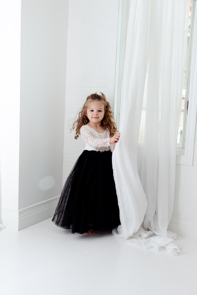 May include: A floor-length dress featuring a white long-sleeved lace bodice and a voluminous black tulle skirt. The waist is accented with a decorative silver-toned embellishment featuring clear crystals, creating a contrast between the lace and skirt.