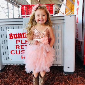 May include: A young girl wearing a pink tulle dress with a sequined bodice and a rose-coloured flower on the waist. She is wearing brown cowboy boots and a pink flower headband. The girl is standing in front of a grey storage container.