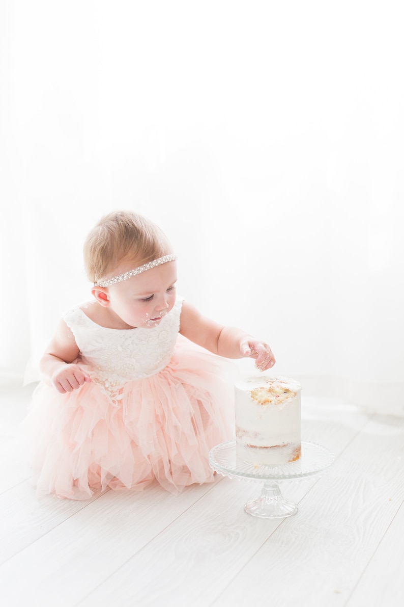 May include: A young girl in a pink tulle dress and a sparkly headband reaches for a white frosted cake with sprinkles on a glass cake stand.