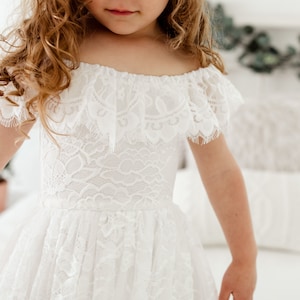 May include: A white off-the-shoulder lace dress with a ruffled neckline and short sleeves. The dress is made of delicate lace and has a flowing skirt.