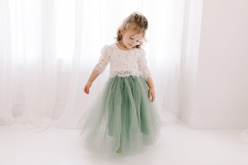 May include: A young girl wearing a white lace top and a sage green tulle skirt. The skirt is long and flowing, and the girl is standing in front of a white background.