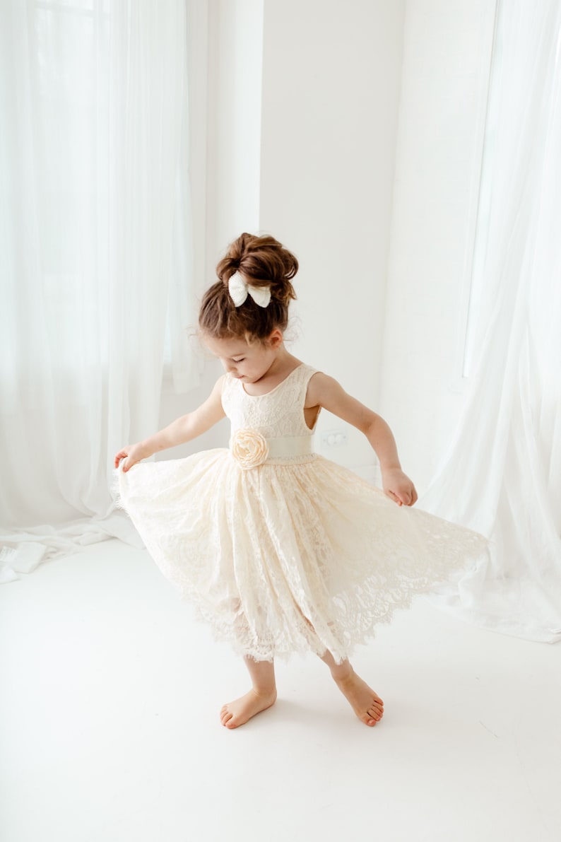 May include: A young girl wearing a white lace dress with a flower accent on the waist. She is twirling in a white room with white curtains.