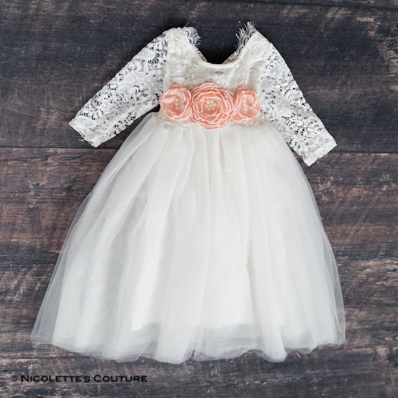 May include: A white tulle dress with long lace sleeves and a peach flower sash. The dress has a scoop neckline and a full skirt.