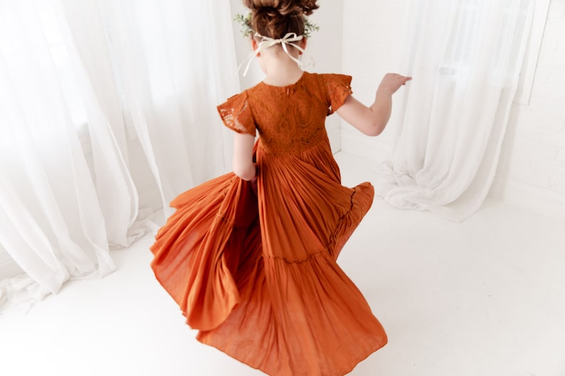 Op de afbeelding: Een jong meisje draagt een roestkleurige gelaagde jurk met kantdetails en korte mouwen. De jurk is zwierig en het meisje draait rond, waardoor een dynamisch beeld ontstaat.