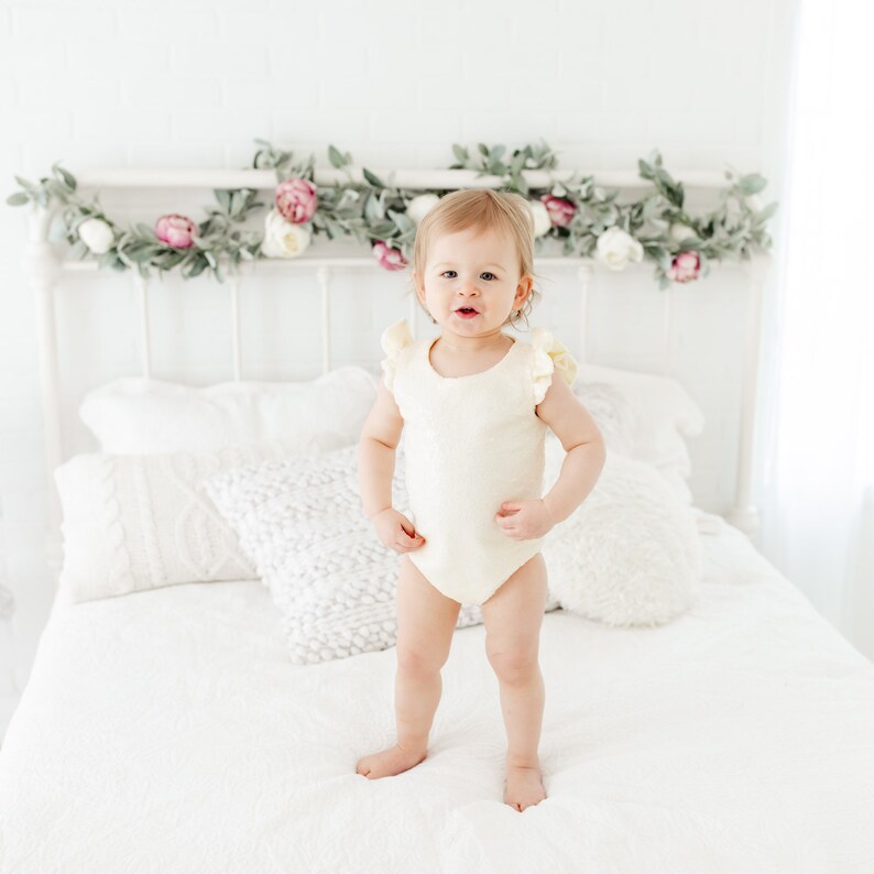 Puede incluir: Un ni&ntilde;o peque&ntilde;o con un body blanco con volantes se para en una cama blanca con almohadas blancas. La cama est&aacute; decorada con una guirnalda de vegetaci&oacute;n y flores rosas.