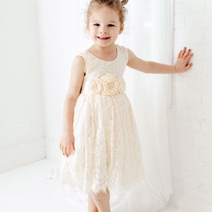 May include: A young girl wearing a white lace dress with a flower sash. She is smiling and standing in front of a white wall.