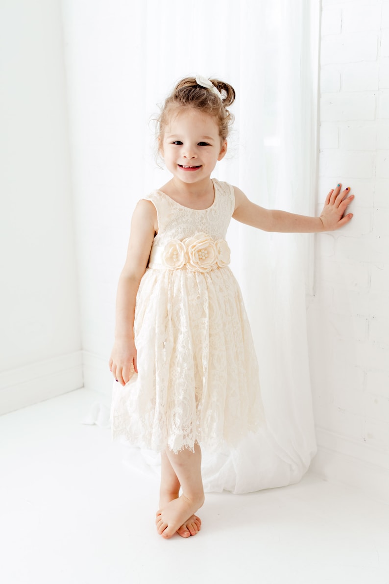 May include: A young girl wearing a white lace dress with a flower sash. She is smiling and standing in front of a white wall.