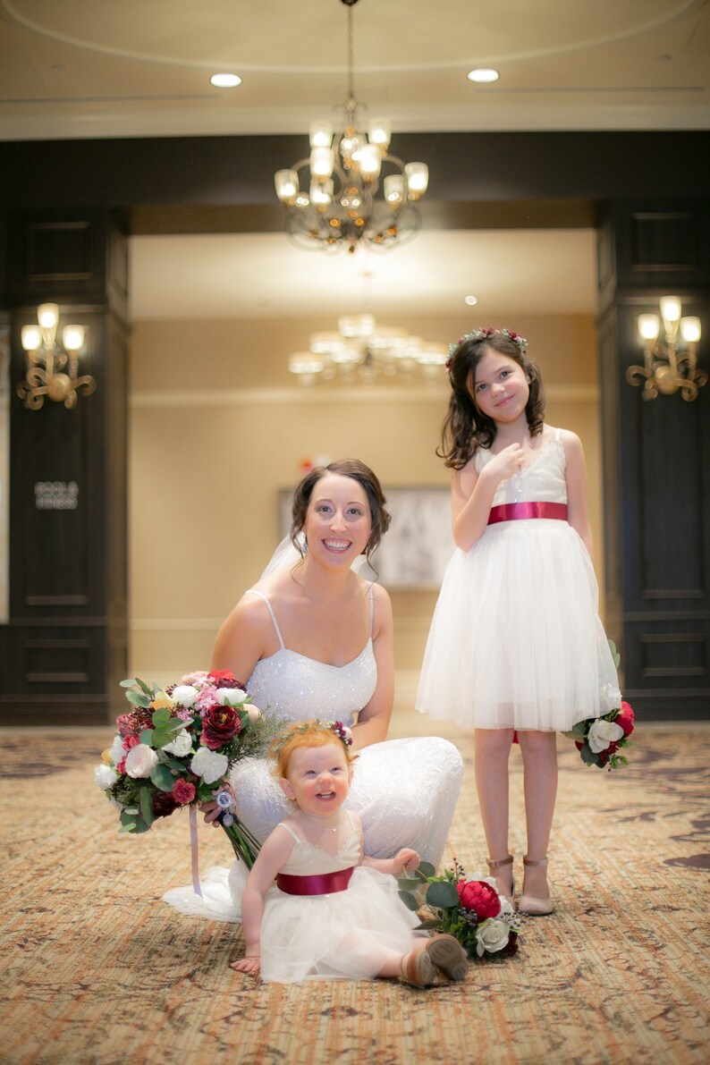 May include: A bride in a white dress with a flower bouquet poses with two flower girls in white dresses. The flower girls are wearing flower crowns and holding bouquets.