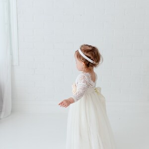 May include: A floor-length dress featuring a white lace bodice with three-quarter length sleeves and a full tulle skirt. The dress includes a large fabric flower accent at the waist and a wide ribbon sash tied into a bow at the back.