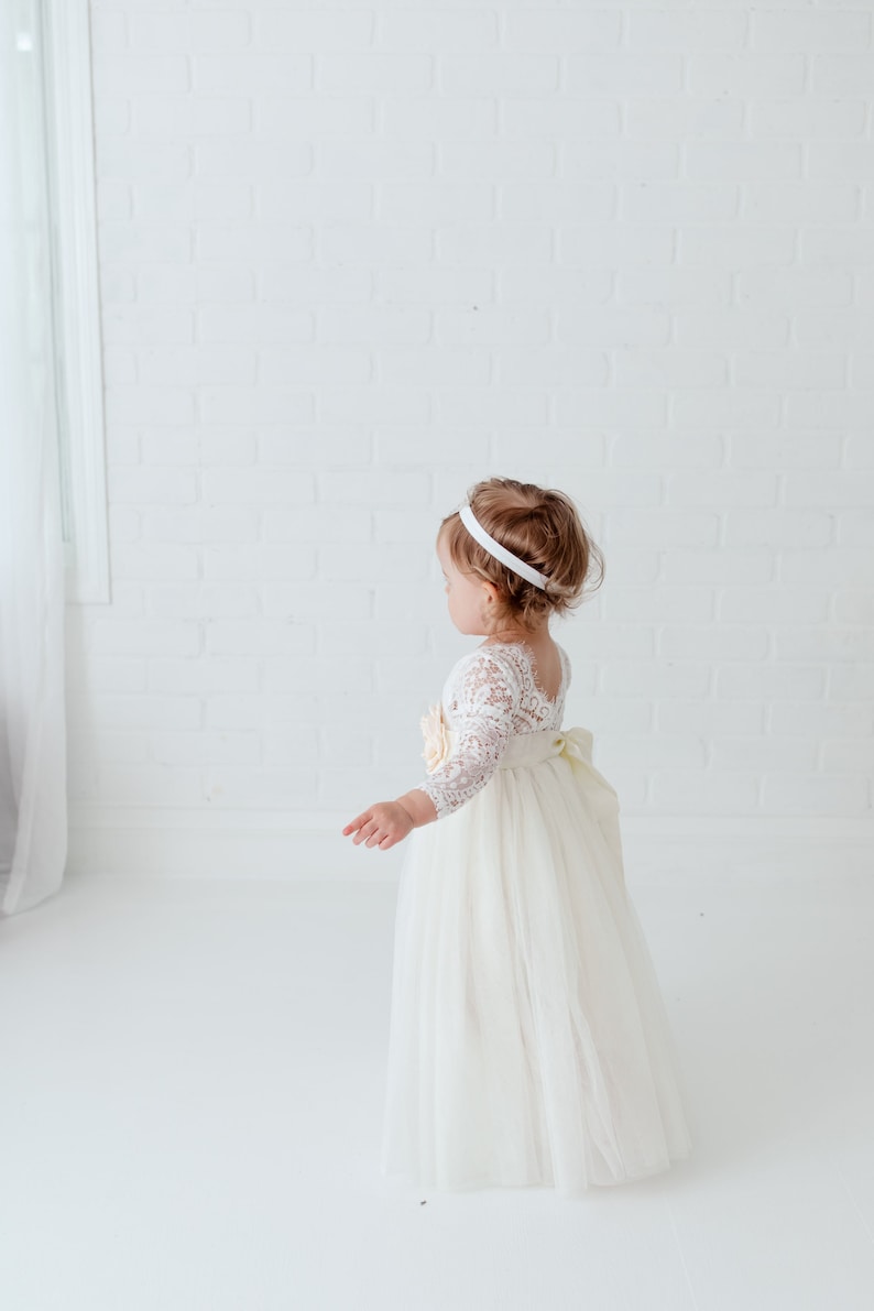 May include: A floor-length dress featuring a white lace bodice with three-quarter length sleeves and a full tulle skirt. The dress includes a large fabric flower accent at the waist and a wide ribbon sash tied into a bow at the back.