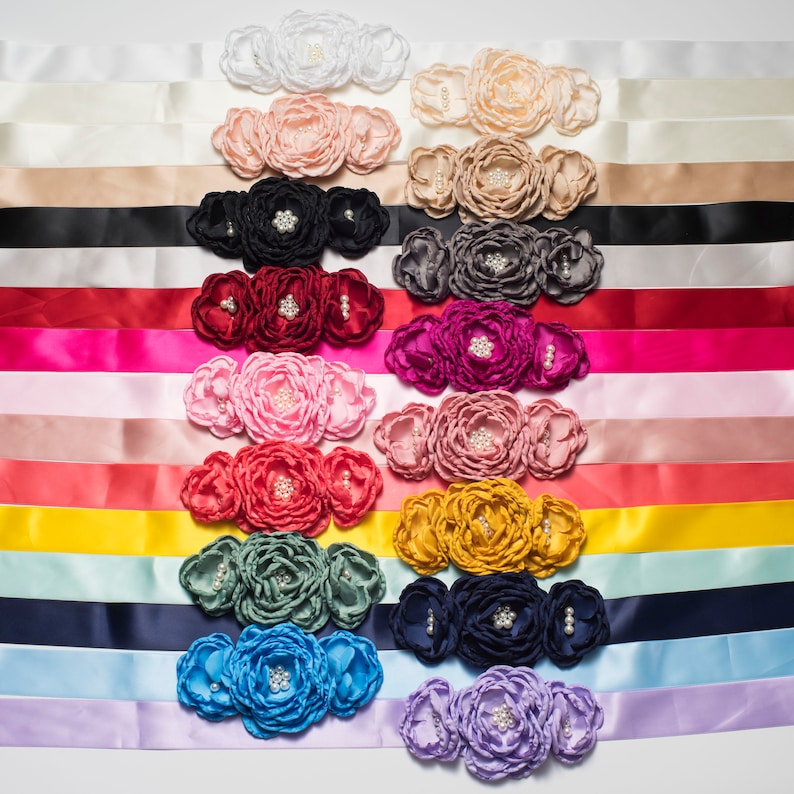Pode incluir: Um conjunto de 12 fitas de cetim em v&aacute;rias cores, cada uma com um enfeite de tr&ecirc;s flores. As flores s&atilde;o feitas de tecido e t&ecirc;m detalhes de p&eacute;rolas.