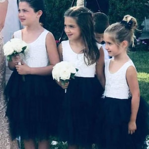 Puede incluir: Tres ni&ntilde;as peque&ntilde;as con vestidos blancos sin mangas, faldas de tul negro y zapatos blancos. Llevan ramos de flores blancas. Las ni&ntilde;as est&aacute;n de pie en un camino de adoquines.