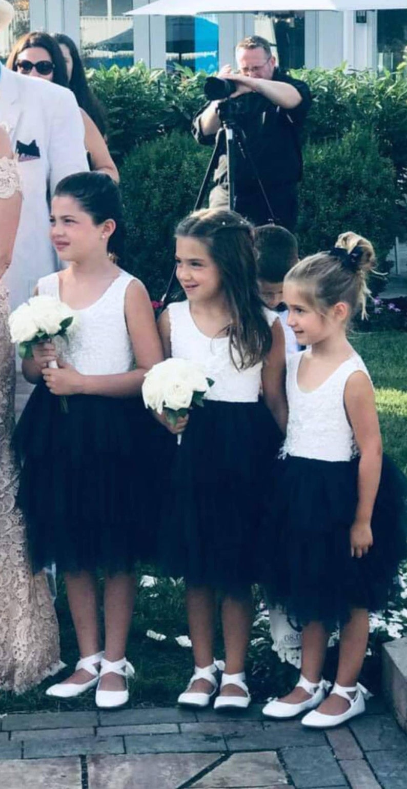Puede incluir: Tres ni&ntilde;as peque&ntilde;as con vestidos blancos sin mangas, faldas de tul negro y zapatos blancos. Llevan ramos de flores blancas. Las ni&ntilde;as est&aacute;n de pie en un camino de adoquines.