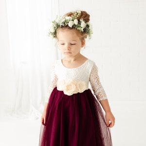 May include: A young girl wearing a white lace dress with a burgundy tulle skirt and a floral crown. The dress has a sash with a large flower embellishment.