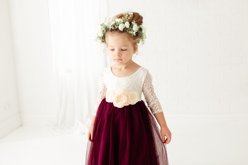 May include: A young girl wearing a white lace dress with a burgundy tulle skirt and a floral crown. The dress has a sash with a large flower embellishment.
