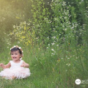 May include: A young girl wearing a white dress and a flower crown sits in a field of green grass and white flowers. She is smiling and looking at the camera.