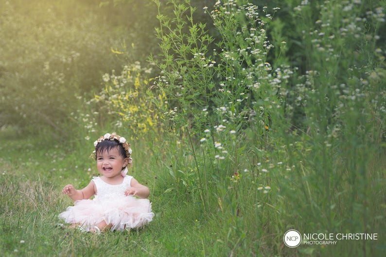 May include: A young girl wearing a white dress and a flower crown sits in a field of green grass and white flowers. She is smiling and looking at the camera.