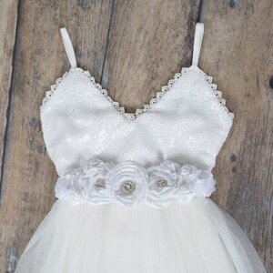 Puede incluir: Vestido blanco de tul con un corpi&ntilde;o de lentejuelas y una faja de flores de sat&eacute;n blanco. El vestido tiene un escote de coraz&oacute;n y tirantes finos.