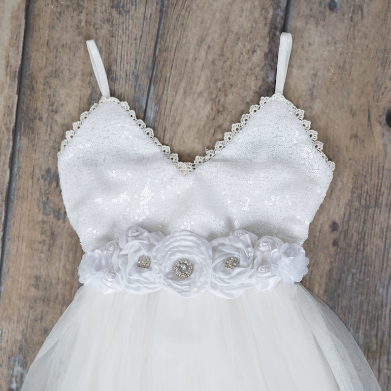 Puede incluir: Vestido blanco de tul con un corpi&ntilde;o de lentejuelas y una faja de flores de sat&eacute;n blanco. El vestido tiene un escote de coraz&oacute;n y tirantes finos.