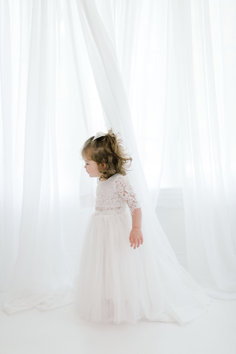 May include: A young girl in a white lace dress with a tulle skirt stands in front of a white curtain.