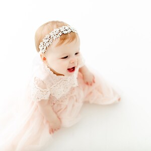 May include: A light pink dress with short lace sleeves and a matching lace bodice, paired with a white floral lace headband. The dress features a layered tulle skirt that flows outward, creating a soft and delicate appearance for this garment.