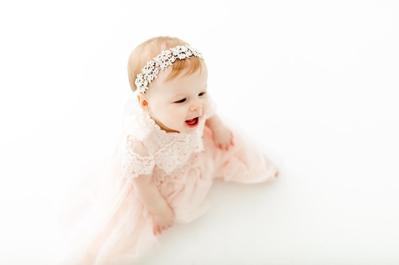 May include: A light pink dress with short lace sleeves and a matching lace bodice, paired with a white floral lace headband. The dress features a layered tulle skirt that flows outward, creating a soft and delicate appearance for this garment.