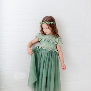 May include: A young girl wearing a sage green tulle dress with a lace bodice and a leafy green crown. The dress has a flowing skirt and the girl is standing on a white background.