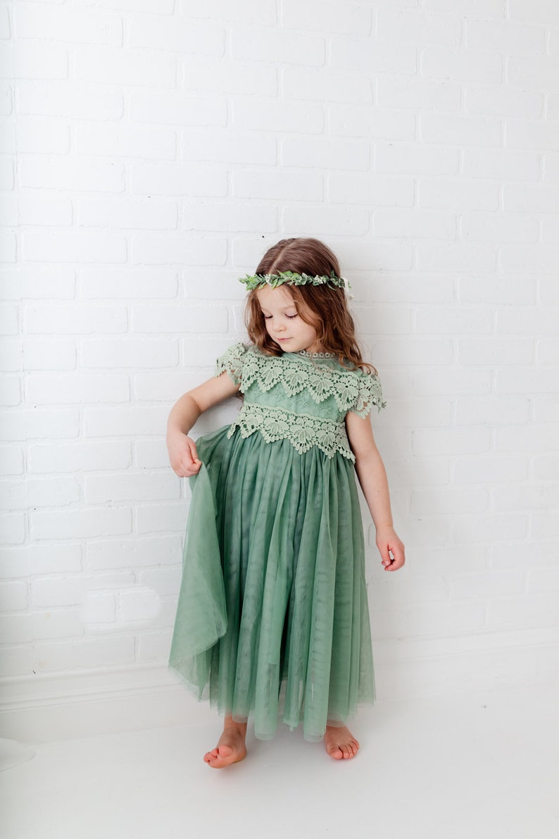 May include: A young girl wearing a sage green tulle dress with a lace bodice and a leafy green crown. The dress has a flowing skirt and the girl is standing on a white background.