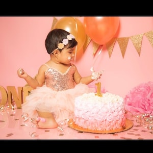 May include: A young girl in a pink and gold sequined dress and a flower headband is celebrating her first birthday. She is sitting in front of a pink cake decorated with pink and white frosting and a gold number one. The cake is on a white plate and there are gold confetti and pink tissue paper balls on the table.