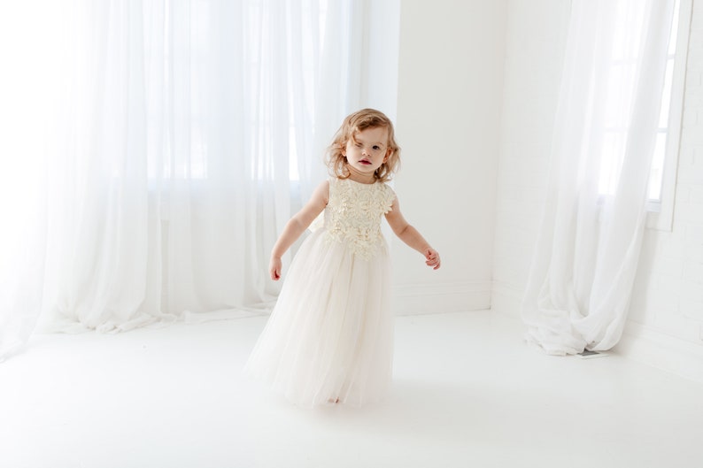 May include: A young girl in a white tulle dress with a floral design on the bodice. She is standing in a white room with white curtains.