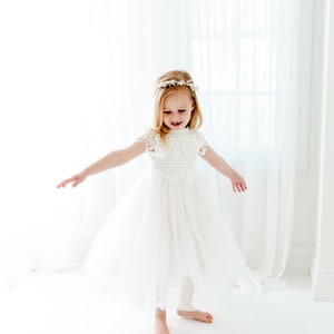 May include: A white dress with a lace bodice, short cap sleeves, and a full tulle skirt. The dress features a high neckline with intricate floral lace detailing. A matching white floral headpiece is worn on the head, completing the ensemble.