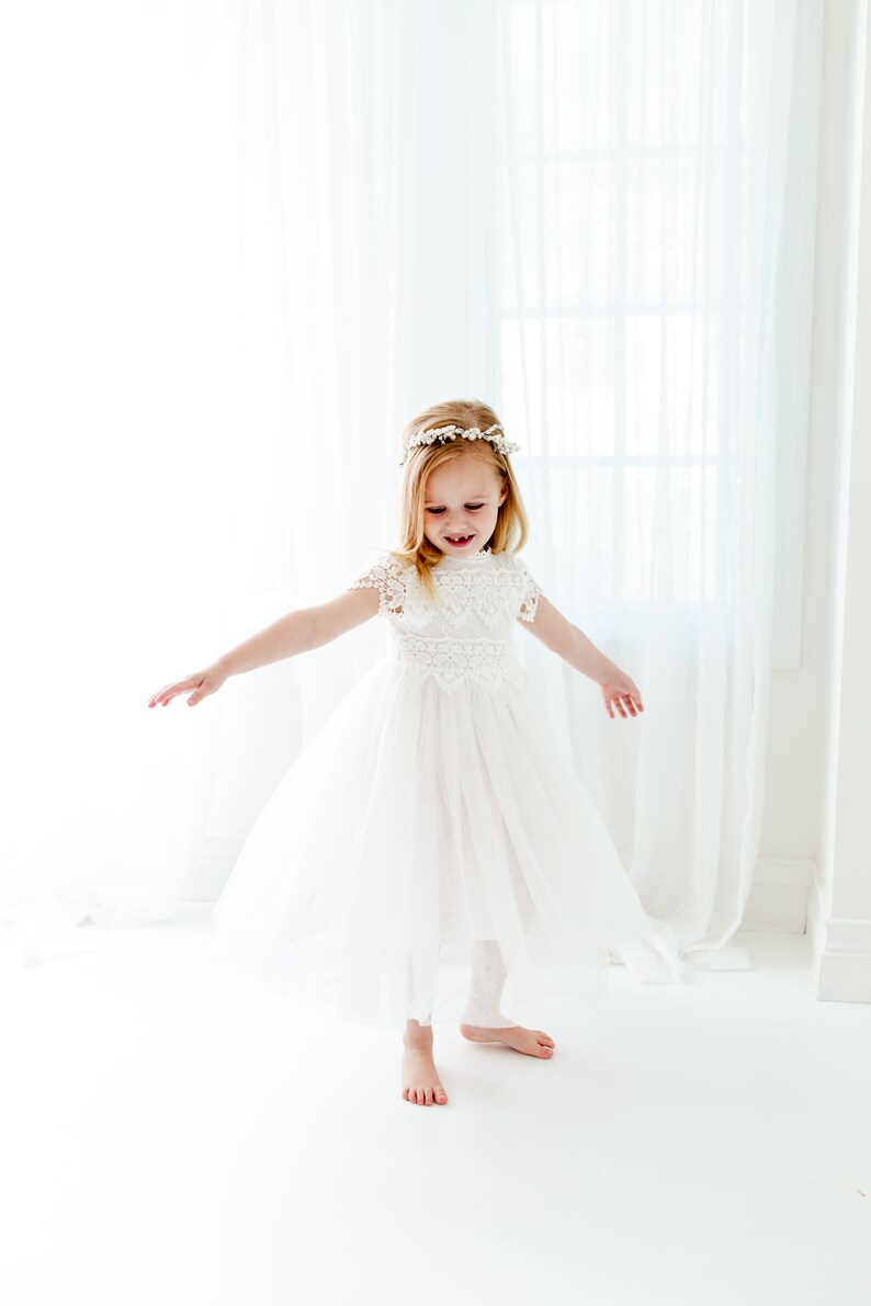 May include: A white dress with a lace bodice, short cap sleeves, and a full tulle skirt. The dress features a high neckline with intricate floral lace detailing. A matching white floral headpiece is worn on the head, completing the ensemble.