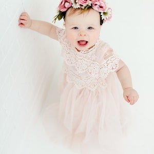 May include: A pale pink dress featuring a delicate lace bodice with intricate floral patterns and a layered tulle skirt. The garment is paired with a floral crown adorned with pink and white fabric flowers and green leaves, worn on the head.