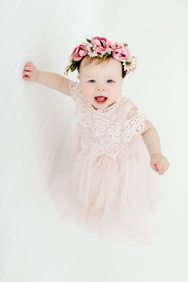 May include: A pale pink dress featuring a delicate lace bodice with intricate floral patterns and a layered tulle skirt. The garment is paired with a floral crown adorned with pink and white fabric flowers and green leaves, worn on the head.