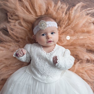 May include: A baby girl wearing a white lace dress and a headband with a rhinestone embellishment lies on a peach-coloured fluffy blanket. The baby is looking up at the camera.