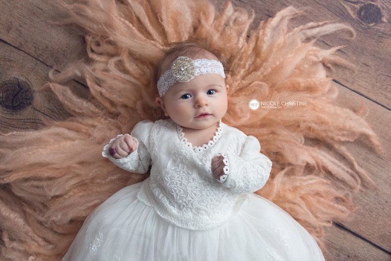 May include: A baby girl wearing a white lace dress and a headband with a rhinestone embellishment lies on a peach-coloured fluffy blanket. The baby is looking up at the camera.