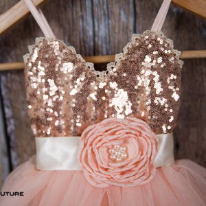 May include: A blush pink tulle dress with a rose gold sequin bodice and a white satin sash. The dress features a large blush pink flower with pearl accents on the front.